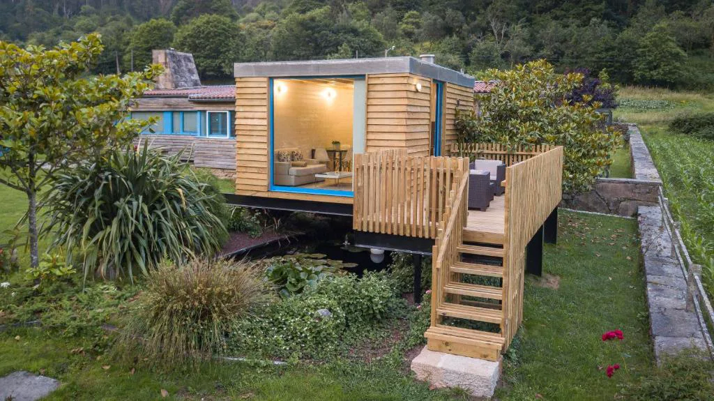 Cabaña de madera elevada sobre un estanque con escalera y terraza iluminada al atardecer, A Cabana de Carmen, Outes