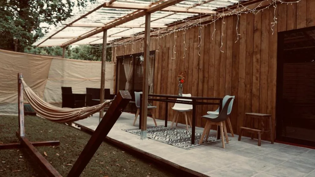 Porche cubierto con hamaca y mesa de comedor junto a la fachada de madera, Cabañas Finca Real, Negreira